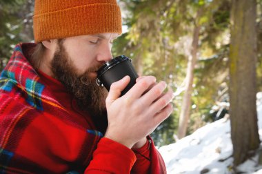 Omuzlarında battaniyeyle kağıt bardaktan içen sakallı adam.