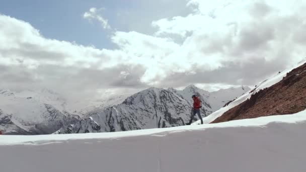 Een jongeman met een pet en rugzak loopt over een besneeuwde bergrug hoog in de bergen. Luchtfoto van romantisch toerisme met besneeuwde toppen en epische wolken — Stockvideo
