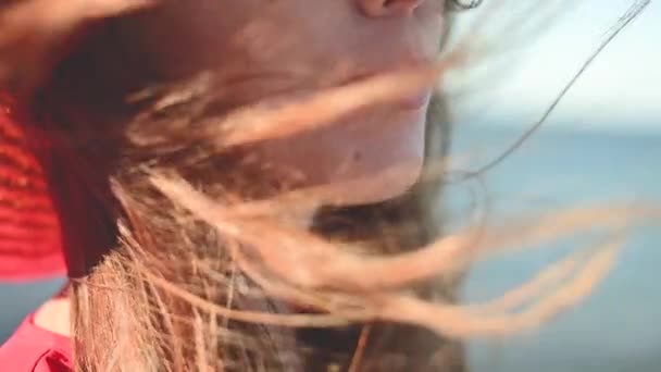 Närbild av en ung vit kvinna hår fladdrar i slow motion i vinden. Grunt skärpedjup. Flicka i röd klänning och hatt drömmare promenad vid havet — Stockvideo