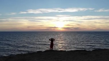 Yalnız, beyaz tenli, kırmızı elbiseli ve hasır şapkalı çekici bir kadının hava manzarası rüzgarda dalgalanan bir uçurumun kenarında, rüzgarlı hava şartlarında duruyor.