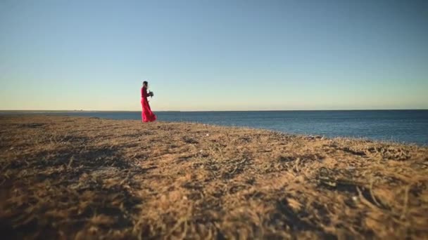 Seorang wanita romantis dengan gaun merah berdiri di pantai tinggi. Sebuah gaun cahaya berkibar-kibar dalam angin. Berpegangan pada topi jerami — Stok Video