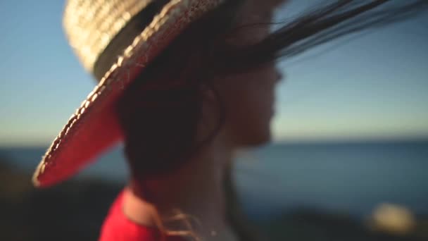 Primo piano di una giovane donna caucasica con i capelli svolazzanti al rallentatore nel vento. Profondità di campo ridotta. Ragazza in abito rosso e cappello sognatore passeggiata al mare — Video Stock