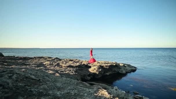 Mujer joven dando una mujer caucásica atractiva delgada en un vestido rojo revoloteando en el viento se encuentra en una roca cerca de la orilla del mar y mira a la distancia. En cámara lenta. Calma y tranquilidad — Vídeos de Stock