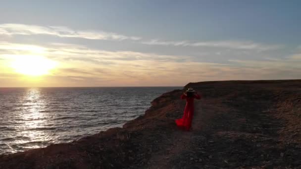Вид с воздуха на одинокую молодую кавказскую привлекательную женщину в красном платье и развевающуюся на ветру соломенную шляпу стоит на краю скалы на закате солнца в ветреную погоду — стоковое видео