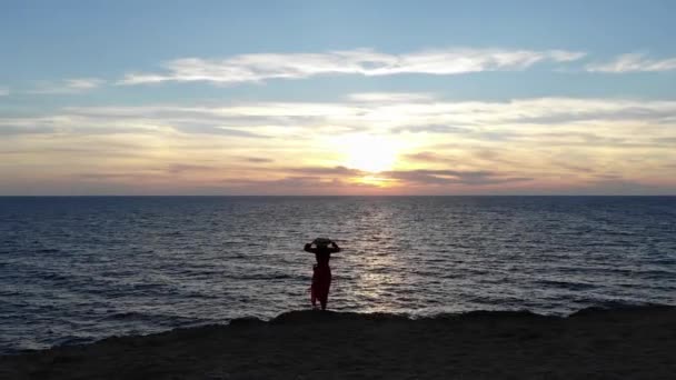 Vista aérea de una joven caucásica solitaria atractiva mujer en un vestido rojo y un sombrero de paja revoloteando en el viento se encuentra en el borde de un acantilado del mar al atardecer durante el tiempo ventoso — Vídeos de Stock