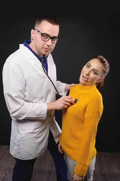 Un jeune médecin à lunettes écoute les battements de cœur d'une femme qui fait des grimaces. Humour médical. Fausses maladies — Photo