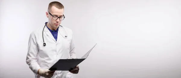 Панорамный портрет привлекательного молодого кавказского врача в очках и белом халате стоит с табличкой в руках, улыбаясь и глядя на табличку на сером фоне. Студийное пространство для копирования — стоковое фото