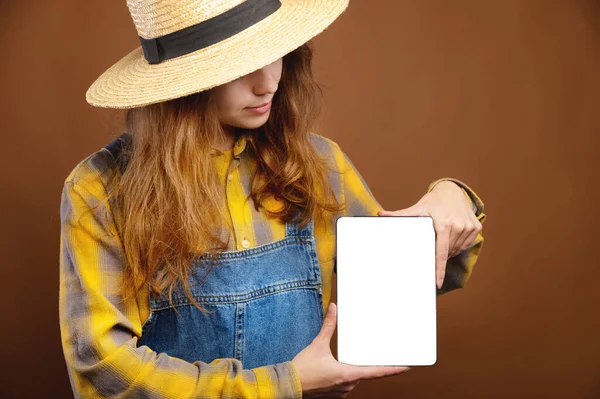 Młoda kaukaska atrakcyjna kobieta z długimi włosami w żółtej koszuli i kapeluszu trzyma tablet i patrzy w ekran gadżet. — Zdjęcie stockowe