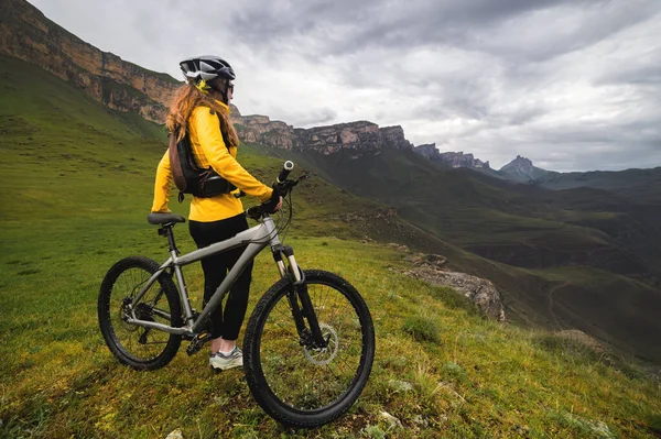 Mujer en una bicicleta de montaña se encuentra en lo alto de las montañas mira a las montañas y el cielo en las nubes. Bicicleta cuesta abajo. Vista trasera —  Fotos de Stock