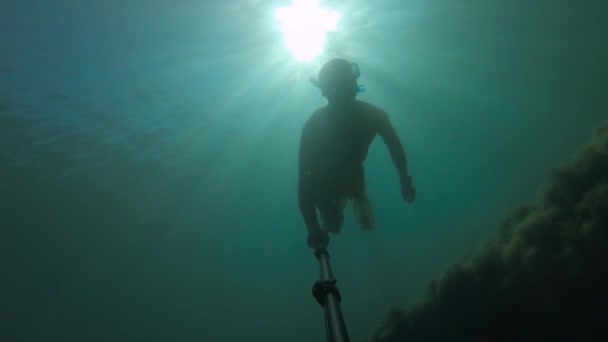 Un joven deportista con una máscara y un snorkel nada con un palo selfie profundo bajo el agua. Deslumbramiento del sol a través del agua — Vídeos de Stock