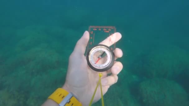 Pandangan orang pertama dari tangan laki-laki memegang kompas magnetik plastik berputar dari samping dan mencari arah yang benar di bawah air. — Stok Video