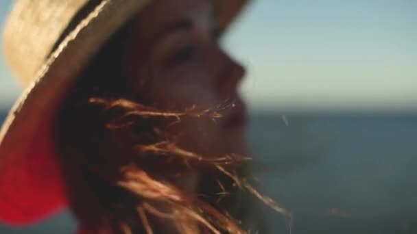 Primer plano de una joven mujer caucásica pelo revoloteando en cámara lenta en el viento. Profundidad de campo superficial. Chica en vestido rojo y sombrero soñador caminar por el mar — Vídeos de Stock
