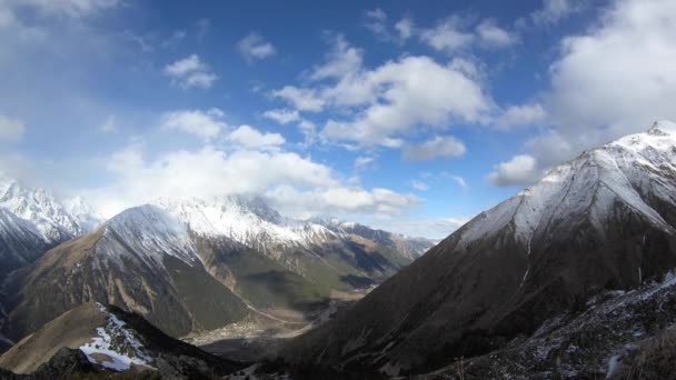 4K timelapse gran angular por encima de la garganta flotan rápidamente nubes que cubren los picos nevados de las altas montañas. El concepto de cambiar el clima en las montañas por debajo del pueblo desfiladero — Vídeos de Stock