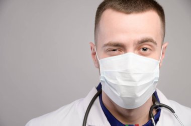 Gri arka planda koruyucu bir maske takmış, dost canlısı, beyaz bir doktorun portresi. Bir adam kameraya bakarak göz kırpıyor ve gülümsüyor.