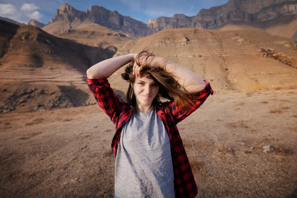 Menina viajante hipster atraente feliz com cabelo ventoso e sorrindo contra o pano de fundo de montanhas ensolaradas copiar espaço. viagens e luxúria. As mãos no cabelo olham para a câmera — Fotografia de Stock