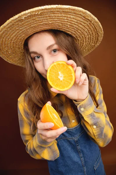 A young attractive Caucasian woman with long hair in denim overalls and a straw hat holds a cut orange in her hands and looks into the camera. Freshness of orange flavor and summer on brown background — Stock Photo, Image
