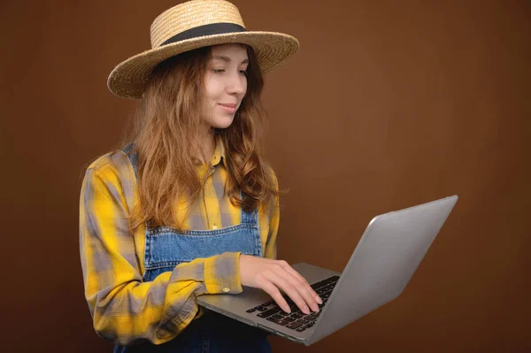 Nevinná atraktivní mladá běloška ve venkovském stylu, oblečená v košili a džínových kombinézách a slaměném klobouku, se podívá na monitor notebooku. Studio portrét. Zemědělská prezentace — Stock fotografie