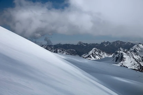 Uma encosta de neve intocada no alto das montanhas do Cáucaso contra o pano de fundo de picos cobertos de neve e nuvens em um dia ensolarado. Campos Freeride em terras montanhosas do planalto — Fotografia de Stock