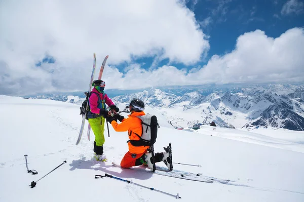 Um jovem esquiador está em seu joelho convidando sua amada namorada, um esquiador, para se tornar sua esposa no alto das montanhas contra o pano de fundo de picos cobertos de neve em um dia ensolarado — Fotografia de Stock