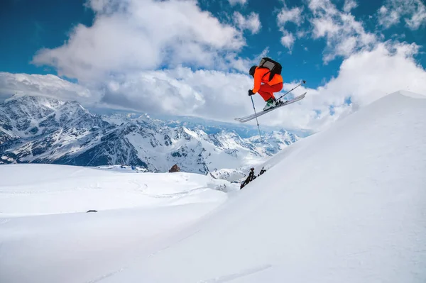 Uno sciatore freerider in tuta arancione con uno zaino congelato in un volo di salto sulle alte cime innevate in montagna in una giornata di sole — Foto Stock