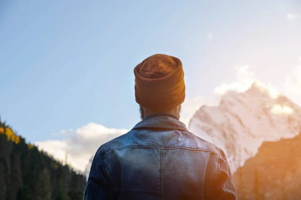 Vista dal retro. Un uomo in giacca di jeans e un cappello marrone si trova nella foresta in montagna sullo sfondo del cielo serale e cime innevate — Foto Stock