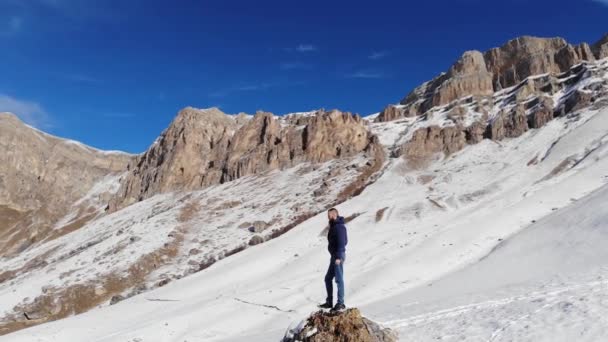 Veduta aerea del giovane uomo caucasico barbuto elegante in piedi su una grande pietra sul pittoresco paesaggio di rocce innevate di montagne. Giorno soleggiato, cielo blu con nuvole — Video Stock