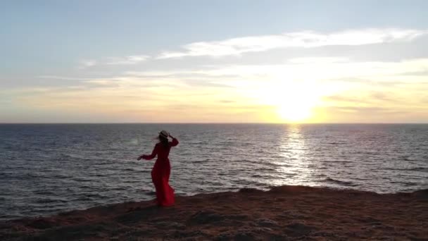 Yalnız, beyaz tenli, kırmızı elbiseli ve hasır şapkalı çekici bir kadının hava manzarası rüzgarda dalgalanan bir uçurumun kenarında, rüzgarlı hava şartlarında duruyor. — Stok video