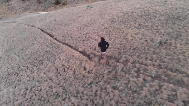 Een luchtfoto van een jong meisje in een pet loopt van een berg heuvel af naar de besneeuwde bergen na zonsondergang in het blauwe uur. Het concept van vreugde in de bergen van wandelen en joggen — Stockvideo