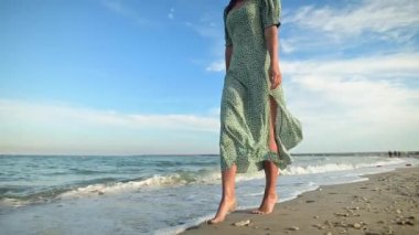 Sabahın erken saatlerinde sahilde yalın ayak yürüyen çekici bir kadın. Altın plajda açık yeşil elbiseli bir turist