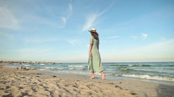 En attraktiv vit smal ung kvinna i lös grön klänning går barfota längs en öde strand nära havet tidigt på morgonen. Långsamma rörelser — Stockvideo