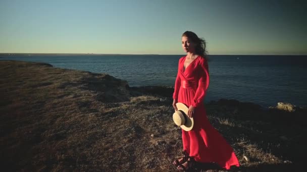 En attraktiv ung vit kvinna i röd klänning med långt hår står med halmhatt i händerna på en stenig havsstrand med slutna ögon i blåsigt väder. Grunt skärpedjup — Stockvideo