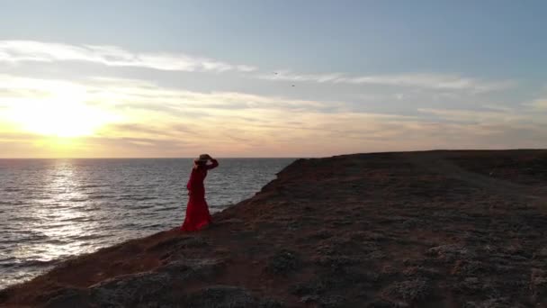 Flygfoto av en ensam ung kaukasisk attraktiv kvinna i en röd klänning och en halmhatt fladdrar i vinden står på kanten av en klippa i havet vid solnedgången under blåsigt väder — Stockvideo