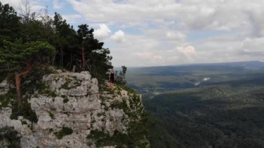 Siyah tişörtlü ve şortlu genç bir adam yüksek bir uçurumun kenarında duruyor ve etrafına bakıyor. Kaya ve ormanlı doğa rezervinin havadan görünüşü