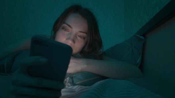 Mujer caucásica joven acostada en una cama en el dormitorio navegando por Internet usando un teléfono inteligente, somnolienta cansada hasta altas horas de la noche. Dramática luz fría y oscura. En Internet, la adicción móvil y el insomnio. — Vídeo de stock