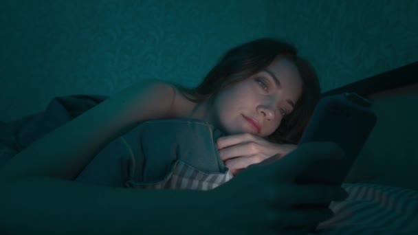 Молодая кавказская женщина лежит в постели в спальне, просматривая интернет с помощью смартфона, сонная, уставшая допоздна. Драматический холодный темный свет. В Интернете, мобильная зависимость и бессонница. — стоковое видео