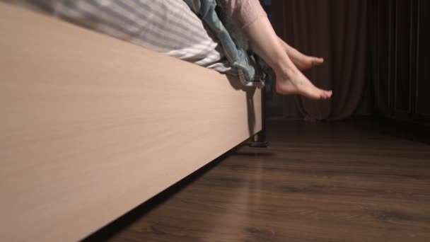 Niedriger Winkel einer jungen kaukasischen Frauenbeine, die vom Bett auf den Boden fallen. Ein Mädchen in Pejamas kommt ans Fenster und öffnet die Vorhänge — Stockvideo