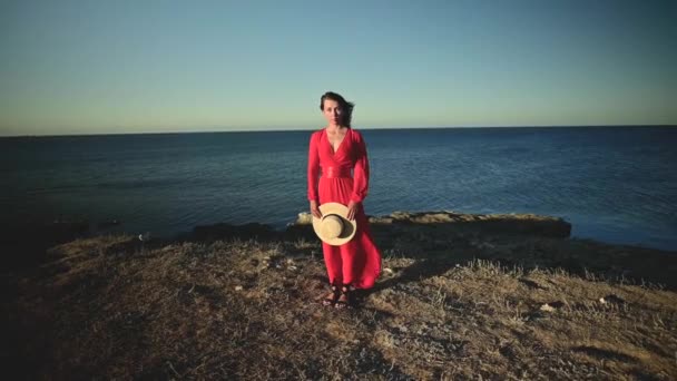 Una atractiva mujer caucásica joven en un vestido rojo con el pelo largo se para con un sombrero de paja en sus manos en una costa rocosa con los ojos cerrados en tiempo ventoso. Profundidad superficial del campo — Vídeos de Stock