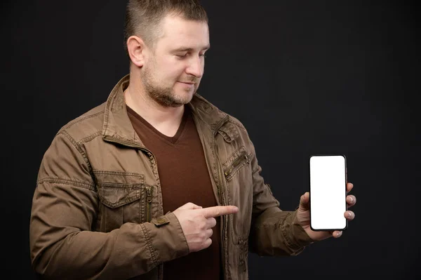 Caucasiano atraente homem maduro em roupas casuais detém um smartphone em suas mãos e aponta para a tela do telefone recortado com o dedo. Corta o ecrã. Estúdio tiro retrato — Fotografia de Stock