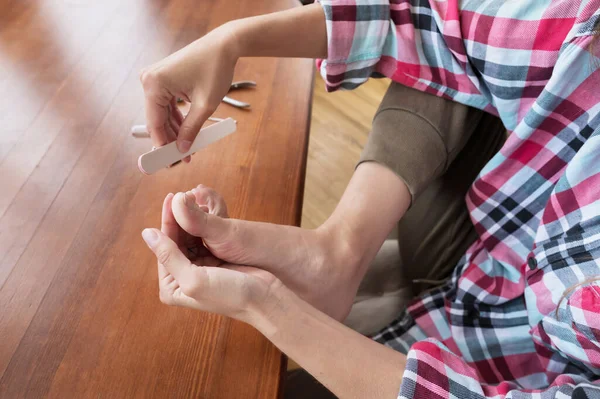 Primer plano de una mujer con ropa casual sentada a la mesa haciendo una pedicura sola en casa — Foto de Stock