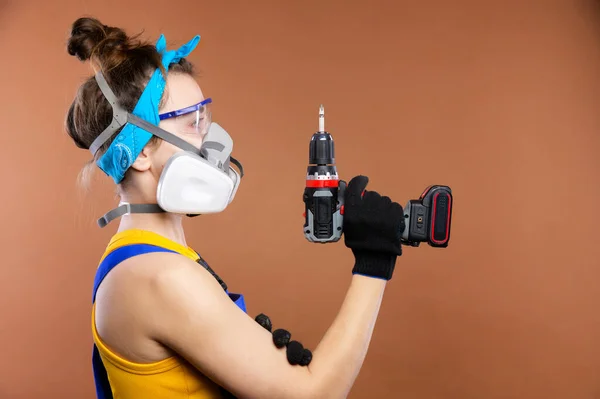 Retrato de estudio de una mujer reparadora con gafas y un respirador con un destornillador eléctrico alimentado por batería. Mujer caucásica carpintero en mono de trabajo — Foto de Stock
