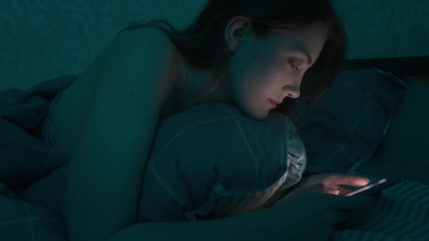 Молодая кавказская женщина лежит в постели в спальне, просматривая интернет с помощью смартфона, сонная, уставшая допоздна. Драматический холодный темный свет. В Интернете, мобильная зависимость и бессонница. — стоковое видео