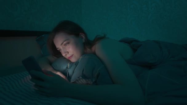 Giovane donna caucasica sdraiata in un letto in camera da letto navigando in Internet utilizzando uno smartphone, stanco sonnolento a tarda notte. Drammatica fredda luce scura. Su Internet, dipendenza mobile e insonnia. — Video Stock