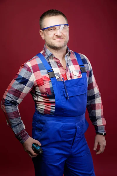 Portret van een positieve vriendelijke blanke mannelijke voorman reparateur in overalls en brillen op een donkerrode achtergrond. Glimlachend in de camera kijkend. Studioshoot. — Stockfoto