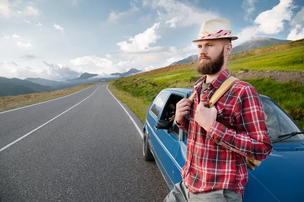 Homem barbudo atraente com uma camisa vermelha xadrez e chapéu com uma mochila. Fica ao lado de um carro retro no fundo de uma estrada pitoresca nas montanhas ao pôr do sol. Viagem fácil, carona — Fotografia de Stock