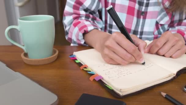 Νεαρή καυκάσια ελκυστική γυναίκα κρατάει σημειώσεις στο σημειωματάριό της ενώ κάθεται στην κουζίνα στο τραπέζι. Χαρτί σημειωματάριο γραφή γυναικεία χέρια ρυθμιστή κίνηση παράλληλη. — Αρχείο Βίντεο