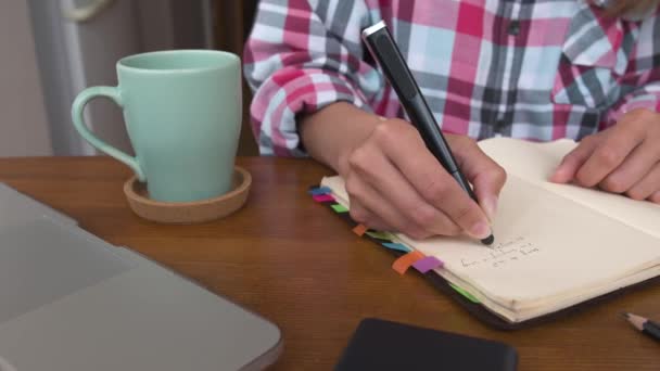 Jonge blanke aantrekkelijke vrouw die aantekeningen maakt in haar notitieboekje terwijl ze in de keuken aan tafel zit. Papier notebook schrijven vrouwelijke handen slider beweging parallel. — Stockvideo