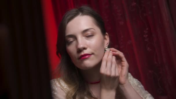Hübsche kaukasische junge Frau kleidet Ohrringe mit Steinen im Schlafzimmer vor dem Schminkspiegel. Boudoir-Fotografie im teuren Rotlicht. Attraktives Mädchen geht zum Ball — Stockvideo