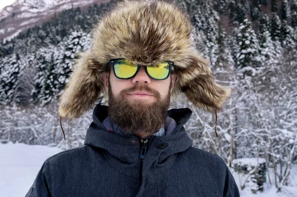 Bonito homem amigo caucasiano em casaco de inverno quente e chapéu de pele e óculos de sol. Olhando para a câmera e sorrindo na floresta de inverno — Fotografia de Stock