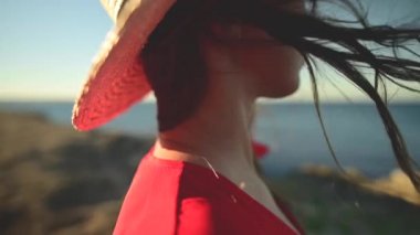 Rüzgarda yavaş çekimde dalgalanan Kafkasyalı genç bir kadının yakın çekimi. Yüzeyin derinliği. Kırmızı elbiseli ve şapkalı kız deniz kenarında yürüyor.