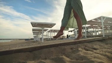 Sabahın erken saatlerinde sahilde çıplak ayakla yürüyen, ağır çekimde ince, çekici bacakları olan bir kadın. Altın plajda açık yeşil elbiseli bir turist